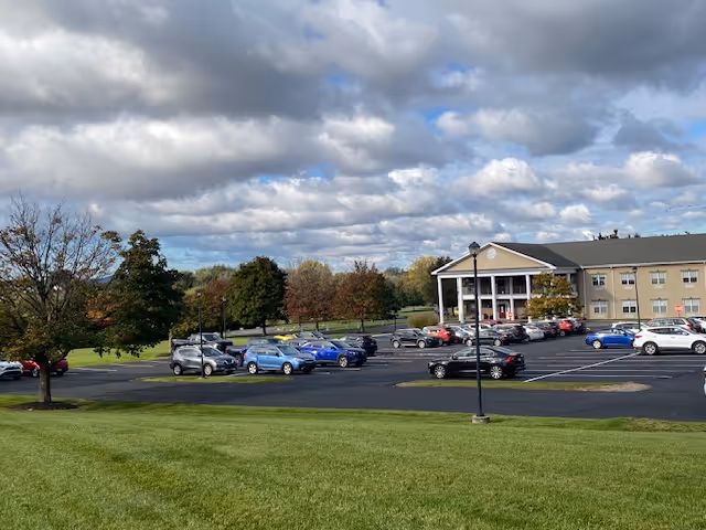 Exterior view of Complete Care at Lehigh Center showing a grassy lawn, parking lot with parked cars, trees, and the facility building under a cloudy sky.