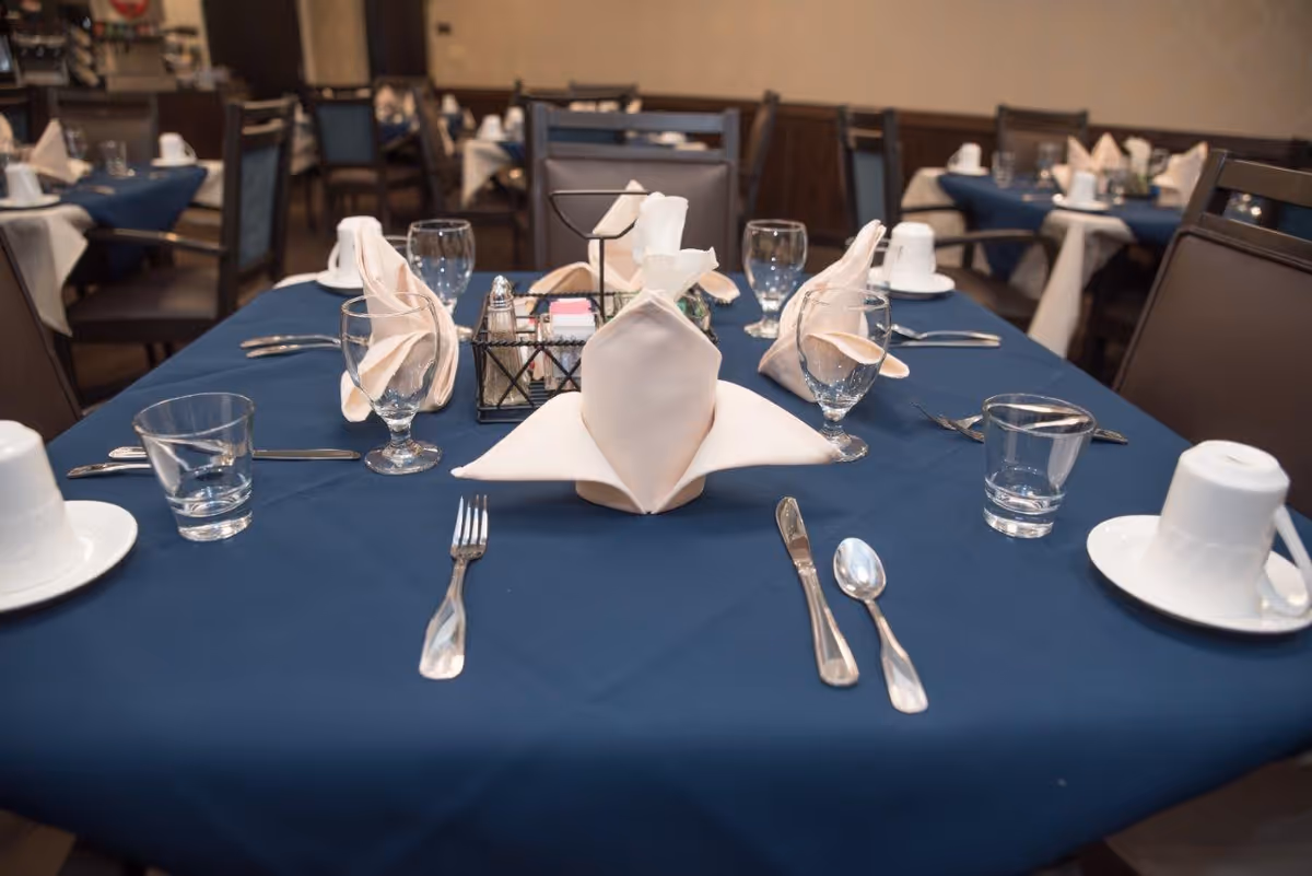 A dining table set with a dark blue tablecloth, neatly folded beige napkins, clear drinking glasses, silverware including forks, knives, and spoons, white coffee cups on saucers, and a condiment holder with salt, pepper, and sugar packets. The background shows more similarly set tables and chairs in a dining room.