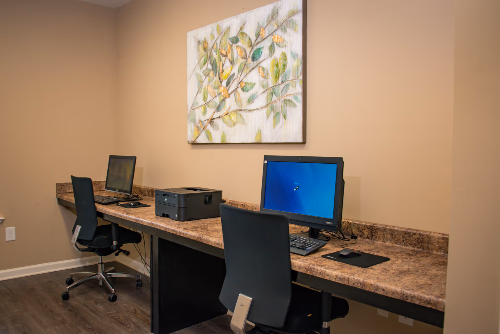A small business-center room with two desktop computers, chairs, a printer on a long counter, and a leaf-themed painting on a beige wall.