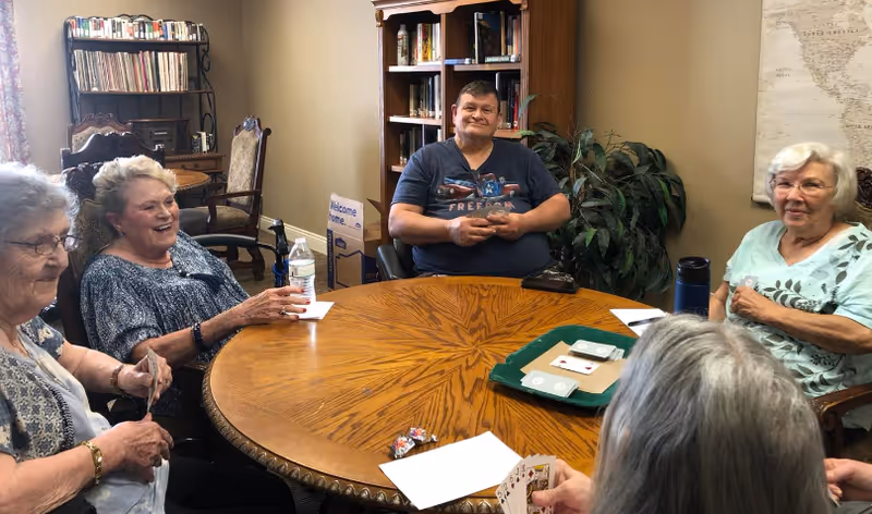 A group of five elderly people sitting around a wooden round table playing cards in a cozy room with bookshelves and a plant in the background. They appear to be enjoying their time together.