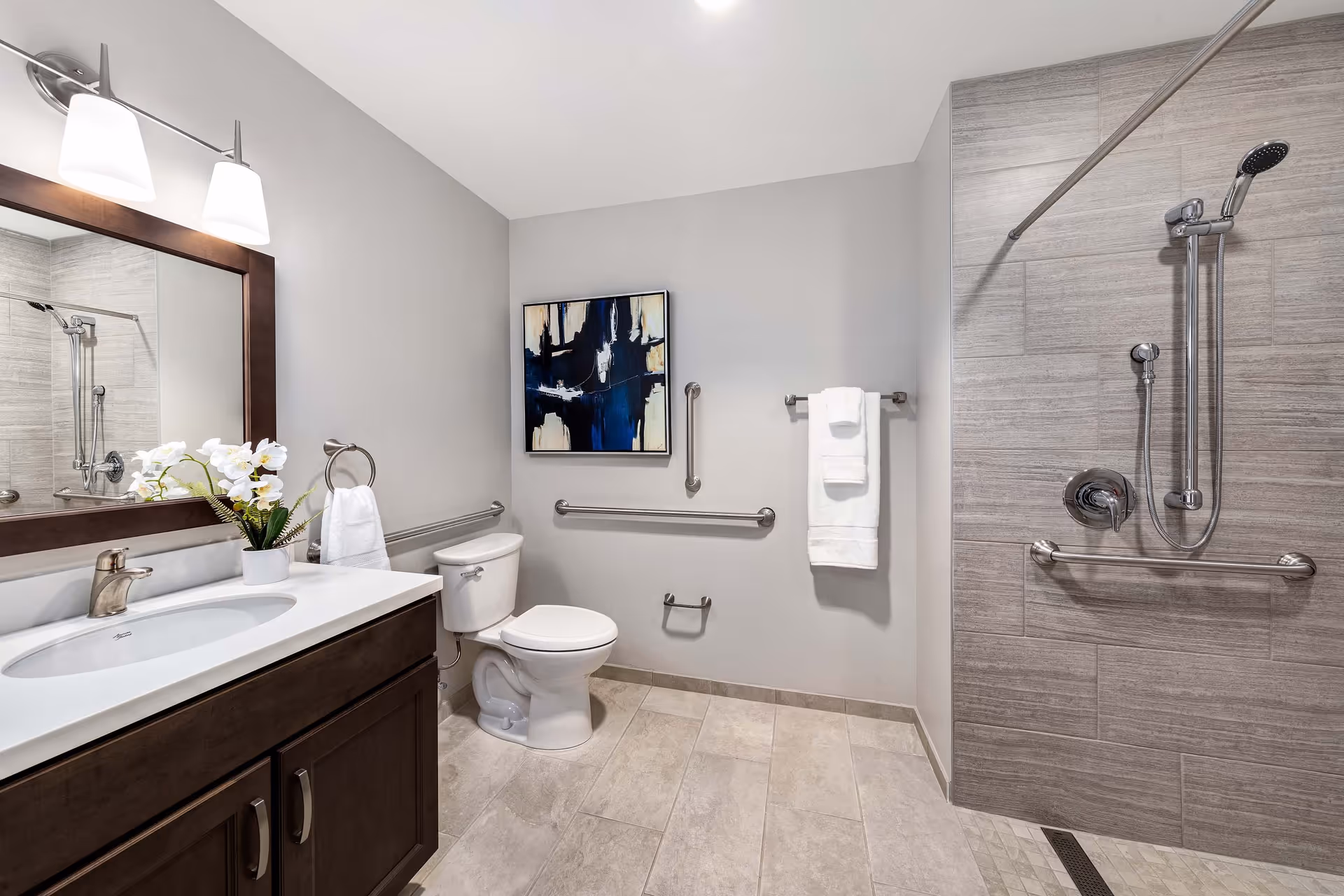A modern bathroom with a white toilet, a dark wood vanity with a white countertop and sink, a large mirror with two light fixtures above it, a walk-in shower with a handheld showerhead and grab bars, and a towel rack with white towels. There is also a piece of abstract art on the wall.