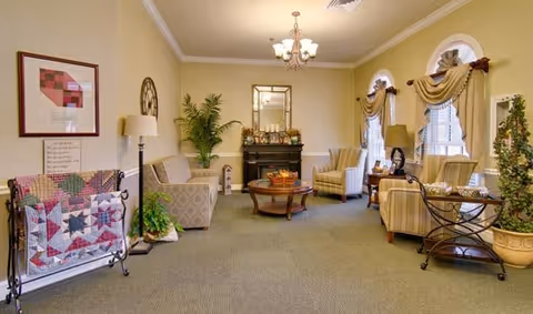 A cozy living room area with beige walls and carpeted floor. The room features a fireplace with a mirror above it, two armchairs, a sofa, a round wooden coffee table with a basket on top, and a side table with a lamp. There are three windows with decorative curtains, a potted plant, a quilt rack with a colorful quilt, and framed artwork on the walls. The room is softly lit by a chandelier and table lamps.
