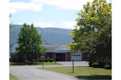 Exterior view of a single-story building with a driveway and garage doors, surrounded by green trees and grass, with mountains visible in the background under a partly cloudy sky.