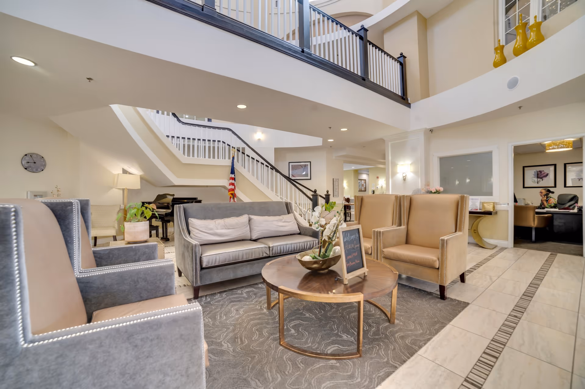 A spacious and well-lit senior living facility lobby with comfortable seating including two gray armchairs, a gray sofa, and two tan armchairs arranged around a round wooden coffee table with a flower arrangement and a small welcome sign. In the background, there is a staircase with white railings, an American flag, a piano, and a reception area with a person sitting at a desk. The floor is tiled with a patterned rug under the seating area, and the walls are painted in neutral tones.