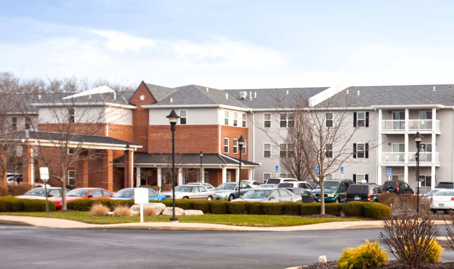 Front exterior of a multi-story senior living building with a parking lot, parked cars, lamp posts, and landscaped grounds.