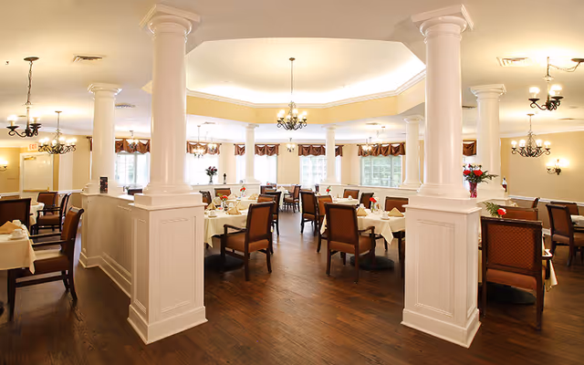 A spacious, well-lit dining room with multiple tables covered in white tablecloths, each set with plates, glasses, and napkins. The room features large white columns, wooden floors, and several chandeliers hanging from the ceiling. Windows with valances allow natural light to fill the space.