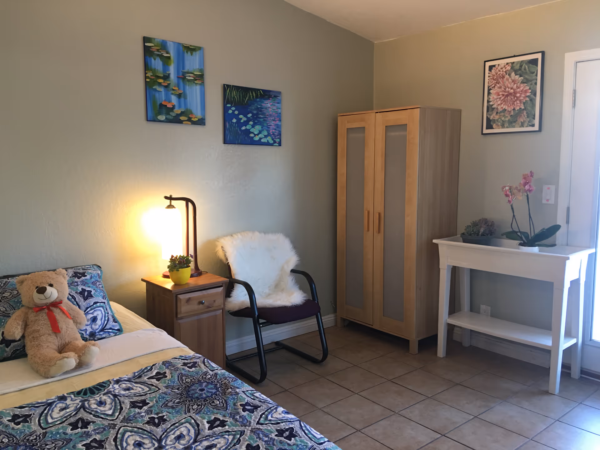 A cozy bedroom with a bed featuring a blue and white patterned bedspread and a teddy bear on the pillow. Next to the bed is a wooden nightstand with a lamp and a small potted plant. A chair with a white fluffy cover is placed against the wall. There is a wooden wardrobe with frosted glass panels and a white console table with two potted plants on top. The walls are decorated with three framed floral paintings. The floor is tiled.