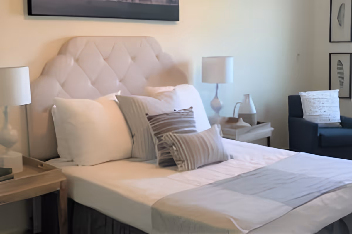 A neatly made bed with a tufted headboard, several pillows in neutral tones, and a light gray throw blanket at the foot. On either side of the bed are wooden nightstands with white lamps and decorative items. In the background, there is a dark blue armchair with a white pillow and framed artwork on the wall.