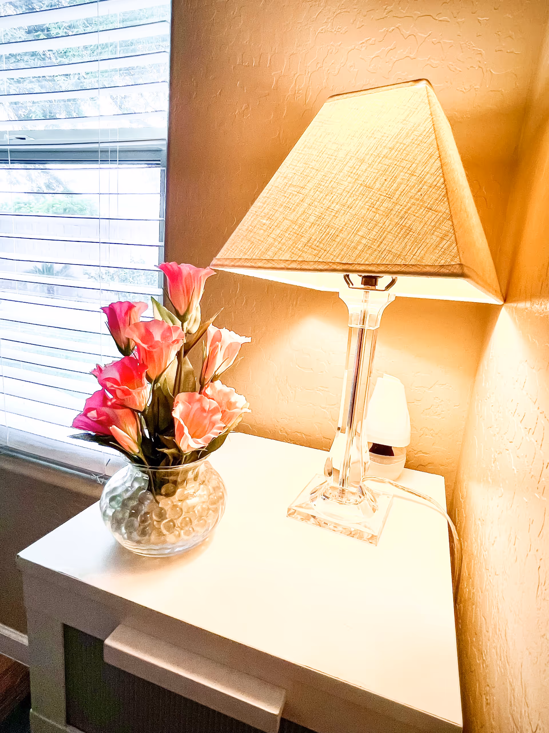 A white bedside table with a clear glass lamp with a beige lampshade turned on, and a round glass vase filled with pink artificial flowers and clear decorative beads. A window with horizontal blinds is partially visible on the left side.