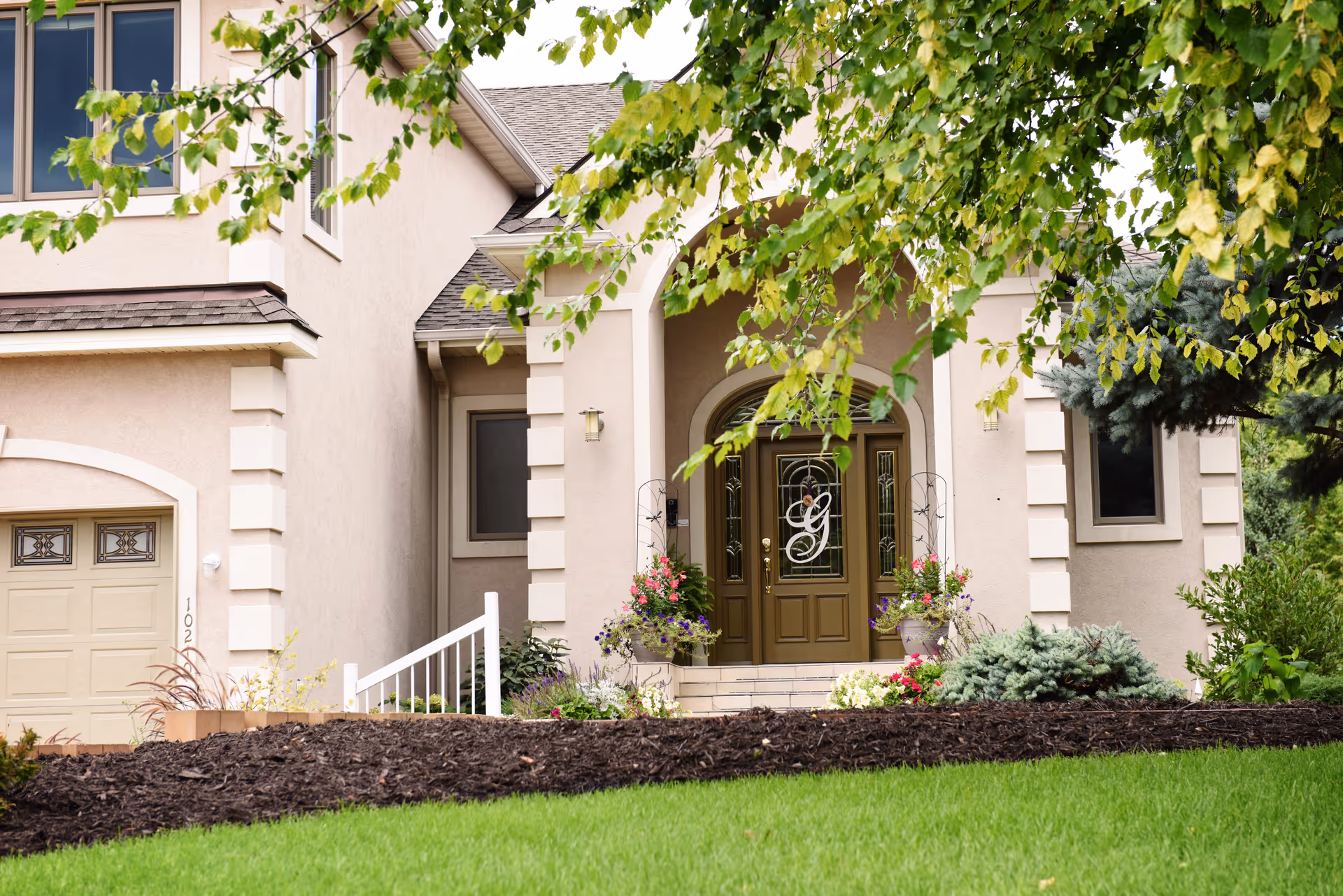 Beige stucco house front with an arched double entry door, potted flowers, garage, and a manicured lawn.