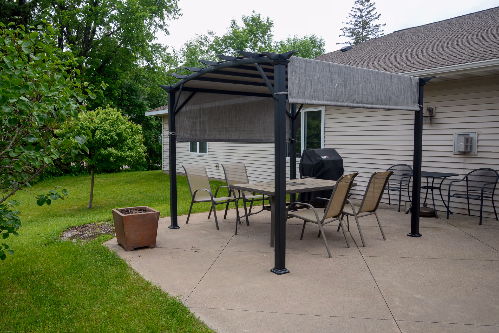 Outdoor patio area with a pergola-style canopy covering a rectangular table surrounded by six chairs. There is a barbecue grill against the beige siding of the building, and additional black metal chairs and a small table are placed nearby. The patio is adjacent to a grassy lawn with trees and shrubs.