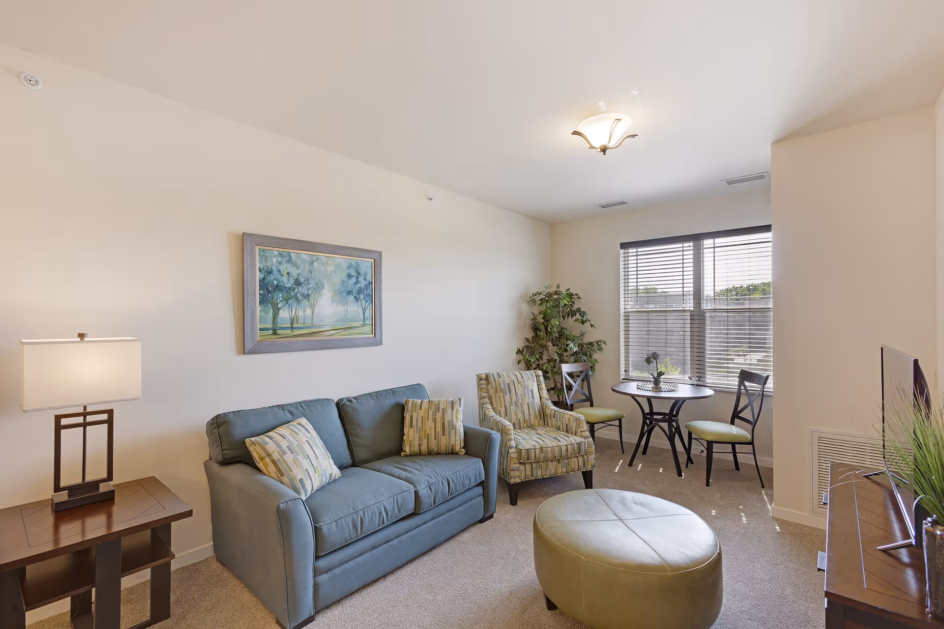 A cozy living room with a blue sofa, two patterned armchairs, a round green ottoman, a wooden side table with a lamp, a small round table with two chairs near a window with blinds, a framed painting of trees on the wall, and a TV on a wooden stand.