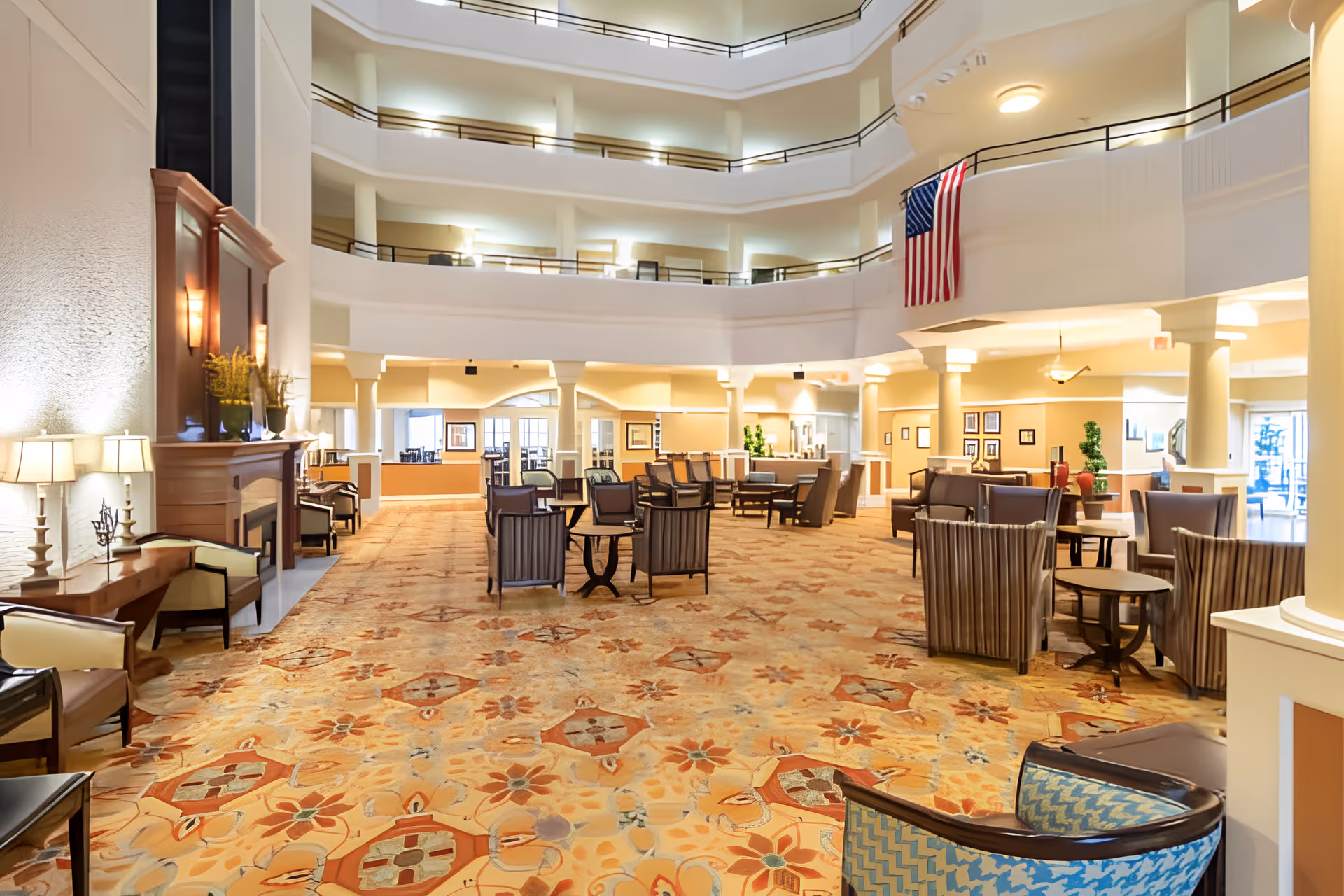 Spacious and well-lit senior living facility common area with multiple seating arrangements including armchairs and small tables, a large patterned carpet, a fireplace with lamps and decorative plants, and an American flag hanging from the balcony above. The area is surrounded by multiple floors with railings overlooking the space.