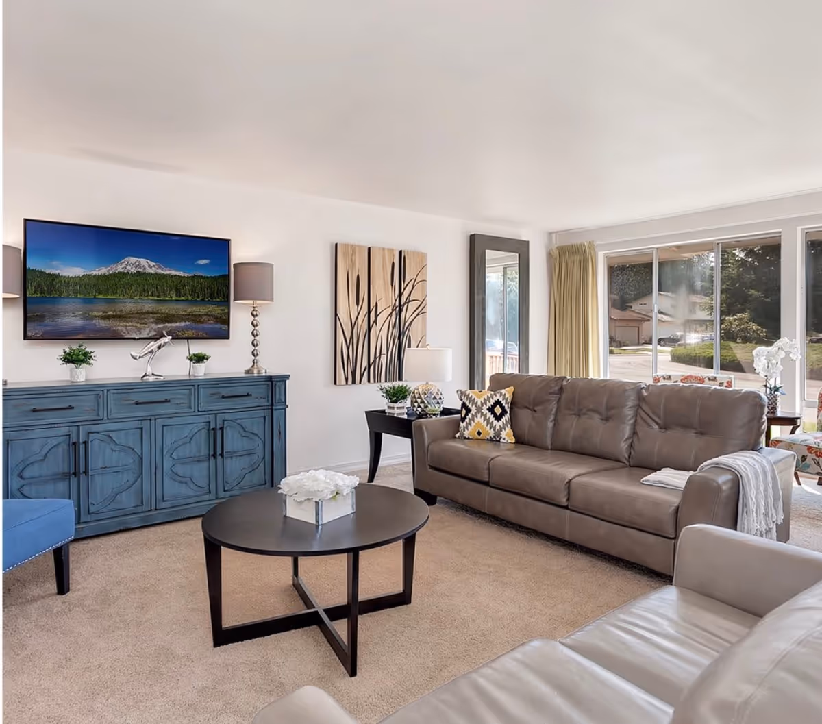 A bright living room with a large brown leather sofa, a round black coffee table with a white flower arrangement, a blue sideboard with decorative items including a lamp and plants, a wall-mounted flat-screen TV displaying a mountain and lake scene, a tall mirror, and large windows with beige curtains letting in natural light.