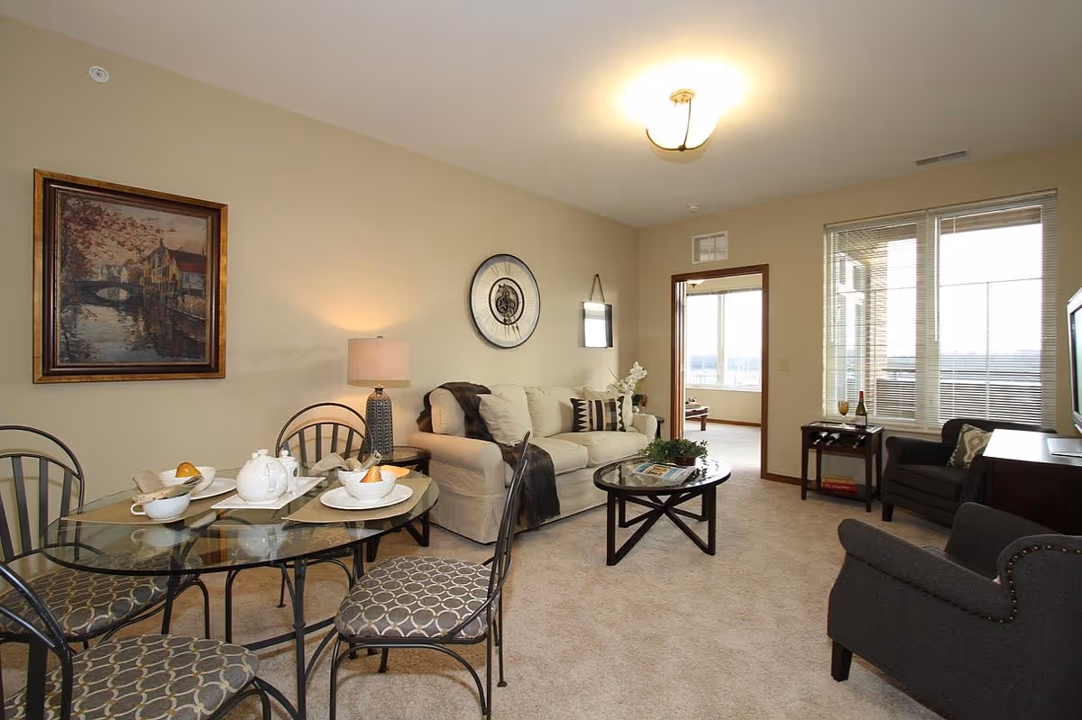 A cozy living room and dining area in a senior living facility named Prairie Gate. The room features a beige sofa with decorative pillows, a glass-top dining table set with white dishes and a teapot, two dark armchairs, a round coffee table with a plant and magazines, a lamp on a side table, a large wall clock, and a framed painting. Large windows with blinds allow natural light to fill the space.