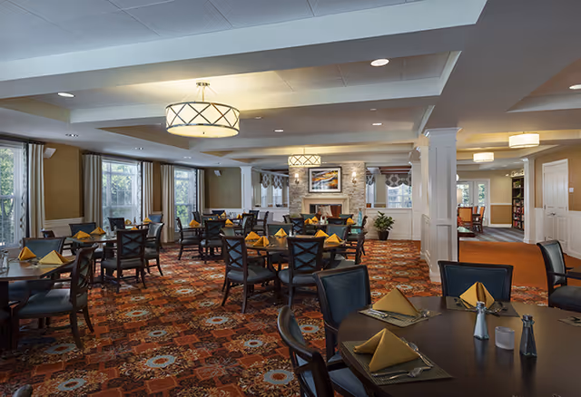 A spacious dining room with multiple round tables set with yellow folded napkins, placemats, and silverware. The room features large windows with curtains, patterned carpet, ceiling lights, and a fireplace with artwork above it in the background.