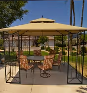 Outdoor seating area with a metal gazebo structure and a yellow canopy. Underneath the gazebo is a round table surrounded by six cushioned chairs with patterned upholstery. The area is set on a concrete patio with green grass and trees in the background, along with a building visible behind the seating area.
