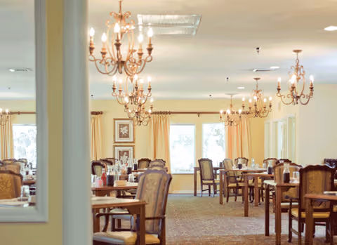 A spacious dining room with multiple wooden tables and cushioned chairs arranged neatly. Elegant chandeliers hang from the ceiling, and large windows with light curtains allow natural light to fill the room.