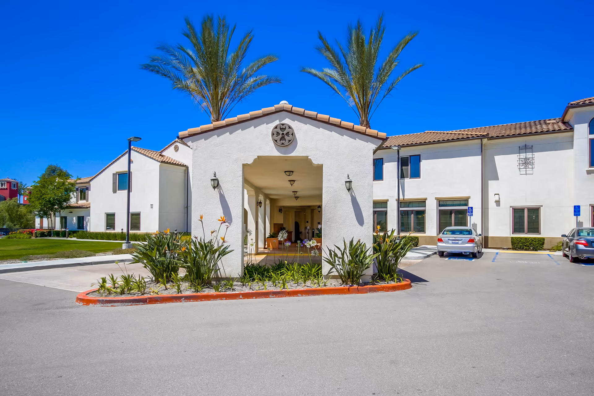 Exterior view of Westmont at San Miguel Ranch facility showing a covered entrance with decorative plants and palm trees, surrounded by a parking area and a two-story building under a clear blue sky.