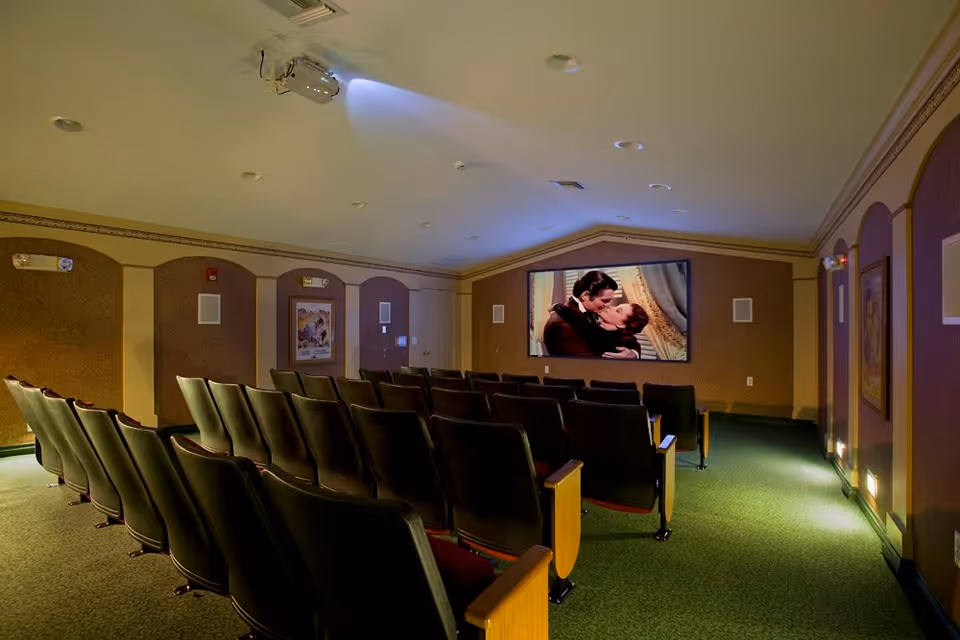 A small movie theater room with rows of black cushioned seats facing a large screen showing a romantic scene from a classic film. The room has a green carpet, beige walls with decorative panels, and a ceiling-mounted projector.