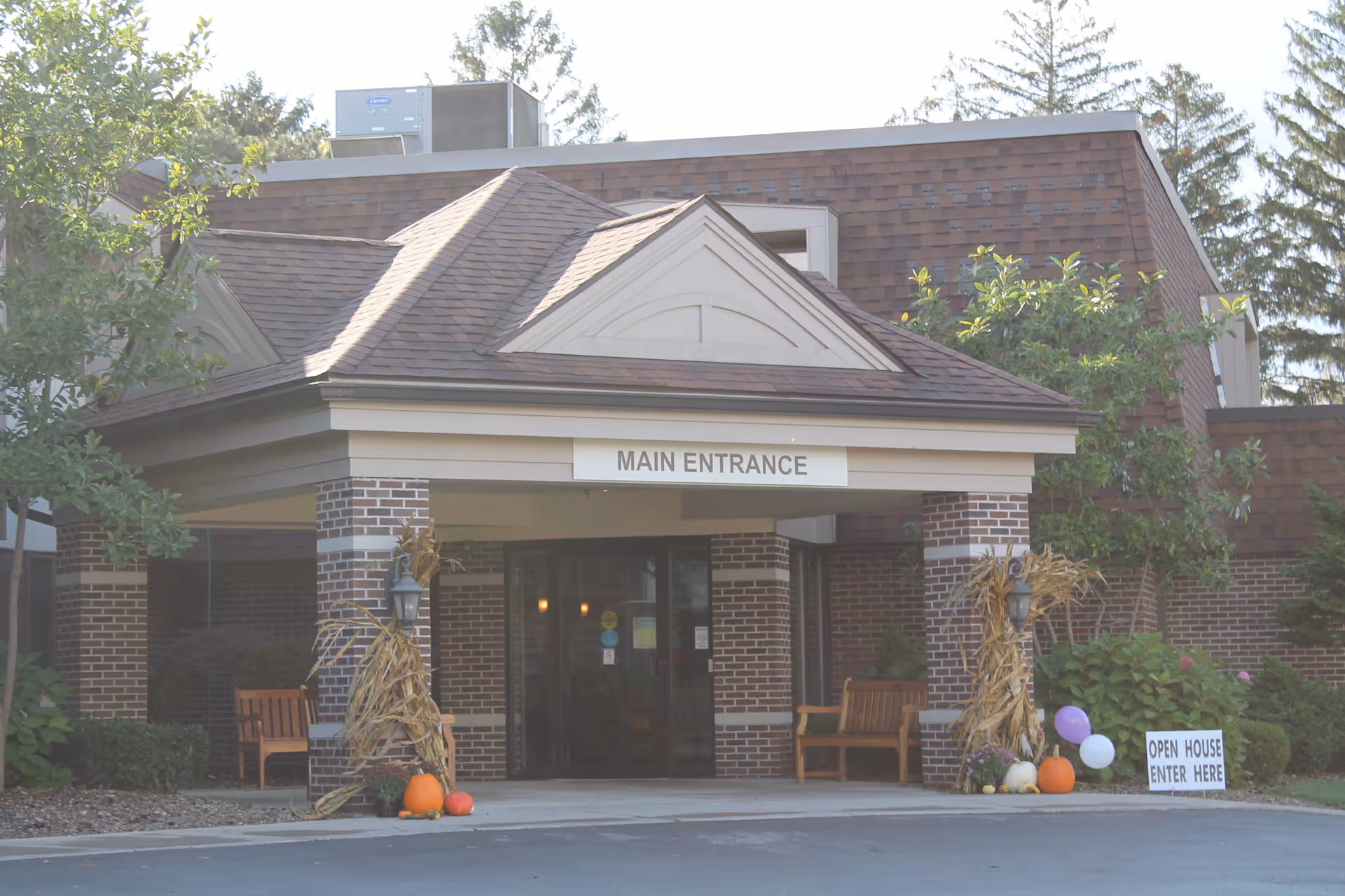 The main entrance of LSS The Good Shepherd Skilled Nursing and Rehabilitation facility, featuring a covered entryway with brick pillars, a sign that reads 'MAIN ENTRANCE', autumn decorations including pumpkins and cornstalks, benches on either side, and a small sign indicating an open house with directions to enter here.