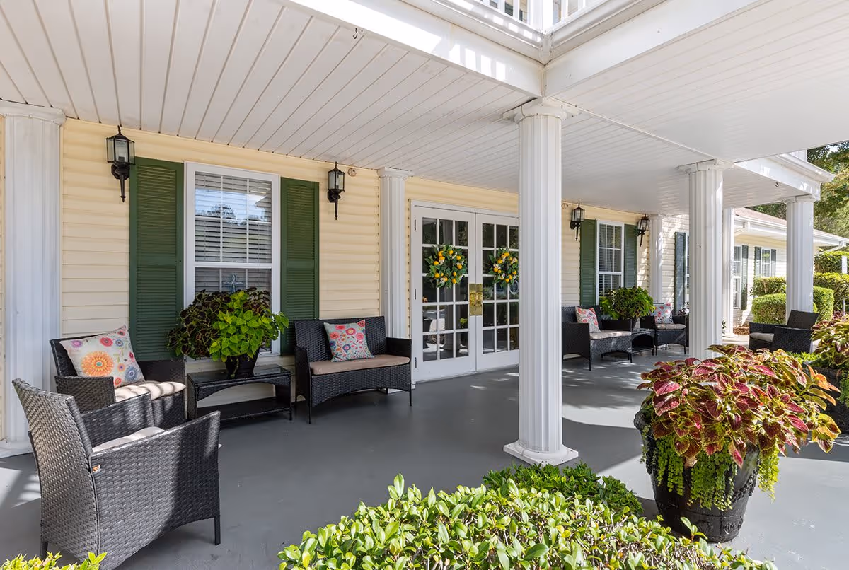 Covered outdoor patio area with white columns and ceiling, featuring black wicker chairs and loveseats with colorful cushions. There are green plants in pots and bushes in the foreground. The building has cream-colored siding with green shutters and double glass doors decorated with wreaths.