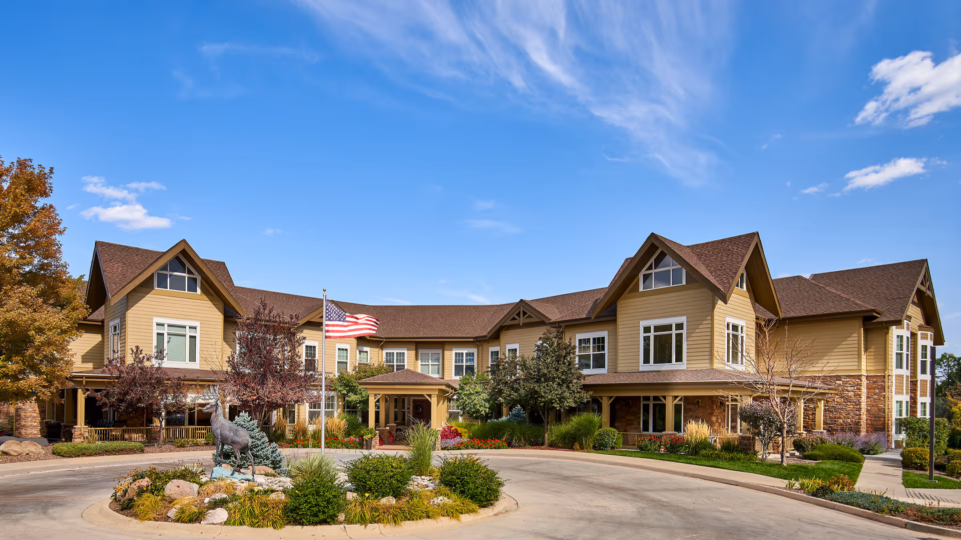 Exterior view of MorningStar Assisted Living & Memory Care of Littleton, a large two-story building with beige siding and brown roof. The entrance is centered with a covered porch, surrounded by landscaped greenery, trees, and a circular driveway with a flagpole displaying the American flag and a deer statue in the middle.
