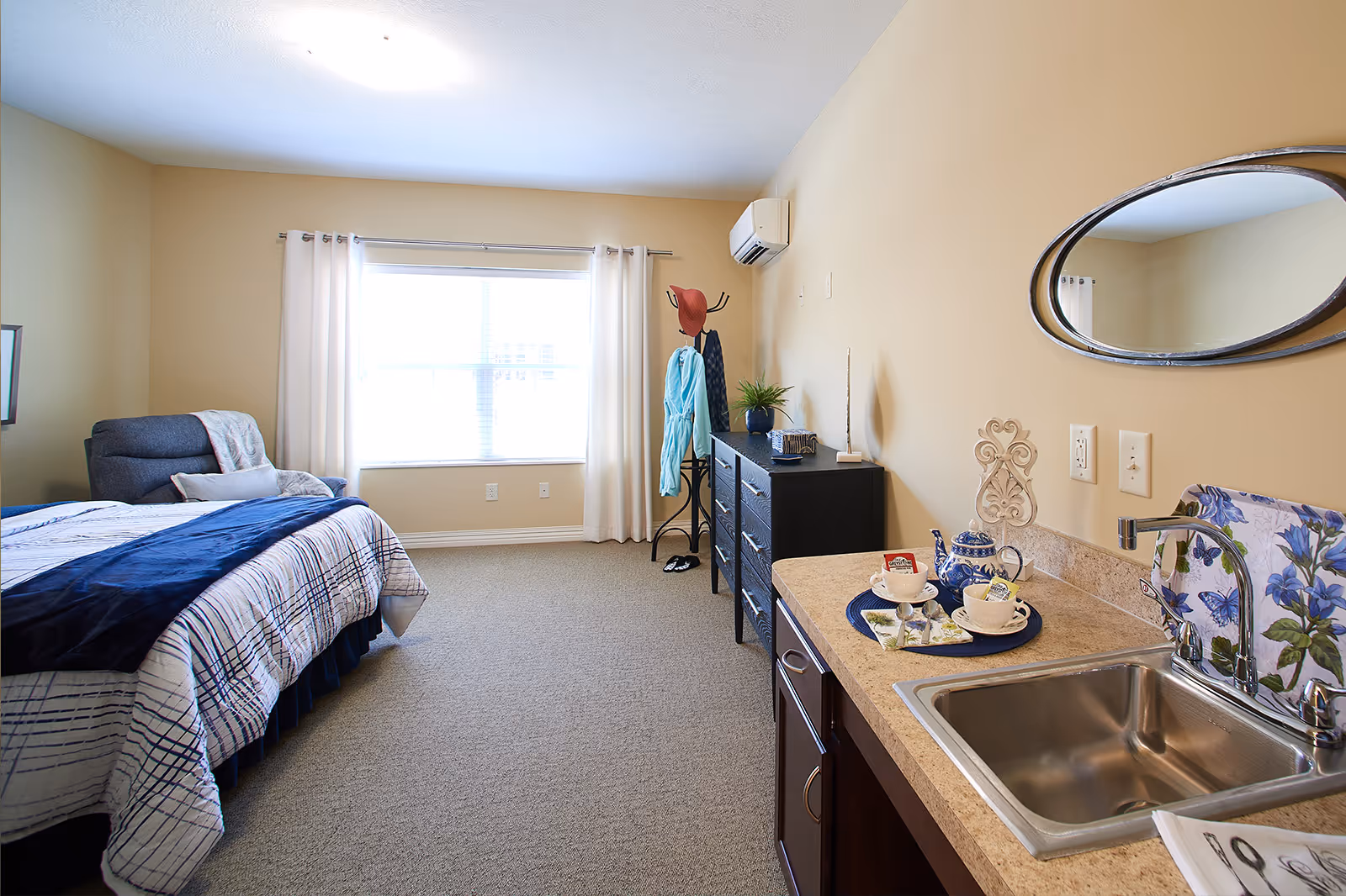A bright, cozy bedroom with a large window covered by white curtains. The room features a bed with a blue and white patterned comforter, a comfortable armchair with a blanket, a black dresser with a plant and decorative items, and a coat rack holding a hat and robe. On the right side, there is a small kitchenette area with a sink, a floral backsplash, and a tray with a teapot and cups. An oval mirror hangs above the kitchenette.