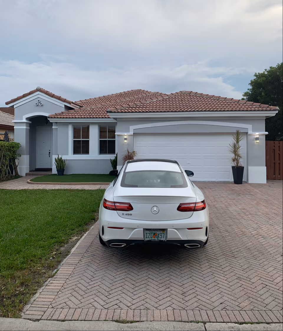 Front exterior view of a single-story house with a tiled roof, a white garage door, and a white Mercedes-Benz E450 parked on the brick driveway. The house has a small front lawn with some plants near the entrance and garage.