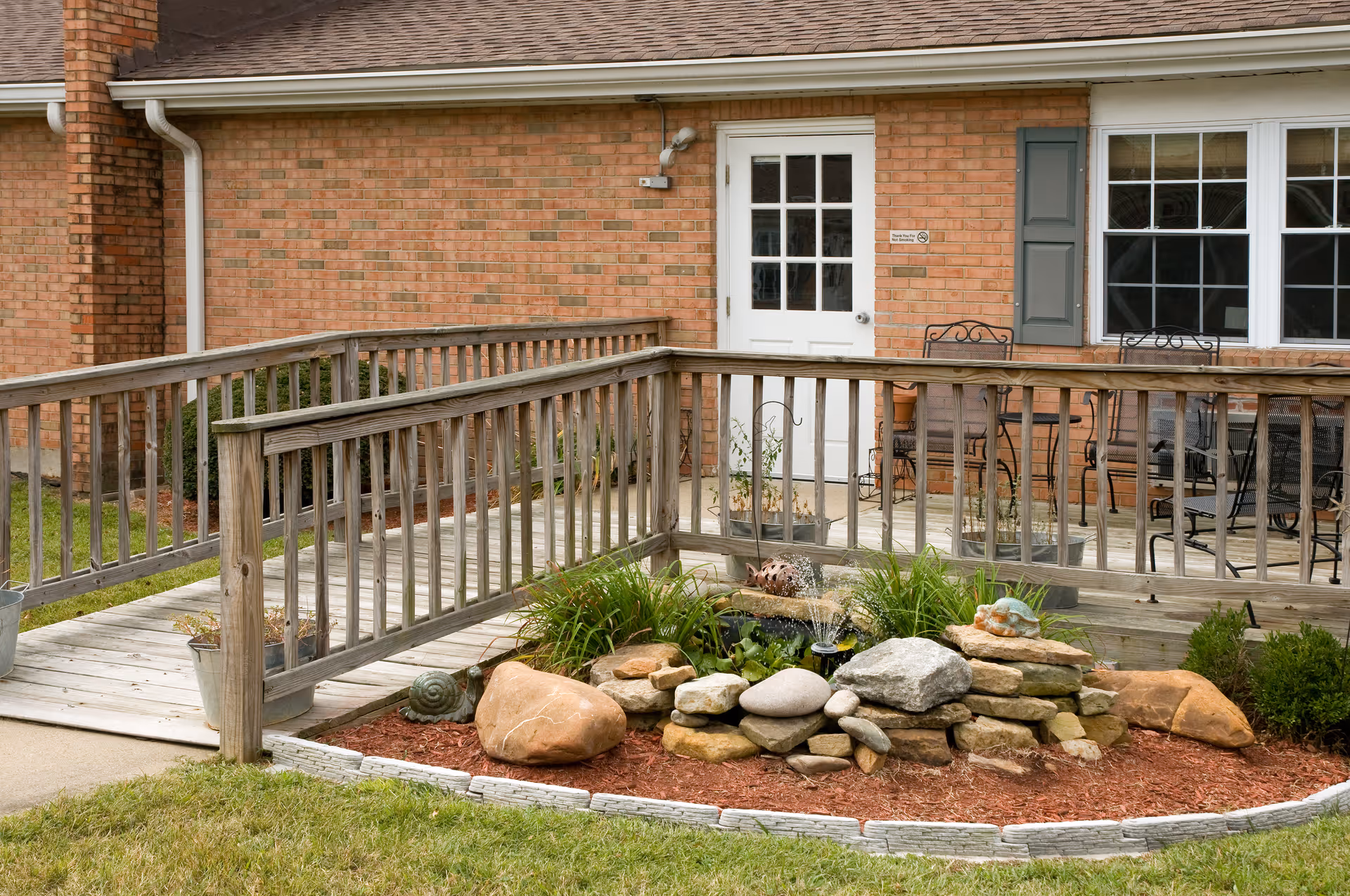 Wooden accessibility ramp and railing leading to a white door and patio with chairs beside a brick building, with a small rock and plant garden in the foreground.