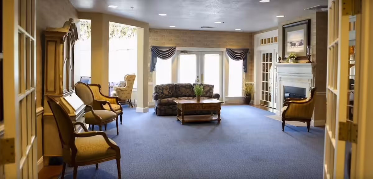 A spacious living room area in a retirement community featuring a blue carpet, a patterned sofa, a wooden coffee table with a plant on it, several upholstered chairs, a fireplace with a framed picture above it, and large windows with drapes allowing natural light to fill the room.