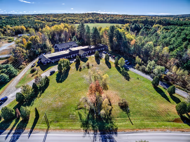 Aerial view of Goudreau’s Retirement Home surrounded by a large grassy area with scattered trees, bordered by dense forest. The building is a single-story structure with a dark roof, situated near a road with a few parked cars visible. The sky is clear with a few clouds.