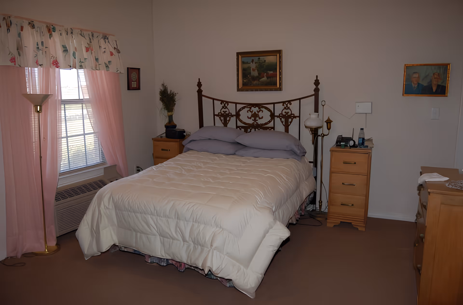 A bedroom with a double bed featuring a decorative metal headboard, beige comforter, and two gray pillows. There are two wooden nightstands on either side of the bed, one with a vase of greenery and the other with various small items. A floor lamp stands near a window with pink curtains and a floral valance. Two framed pictures hang on the walls, and a wooden dresser is partially visible on the right side.