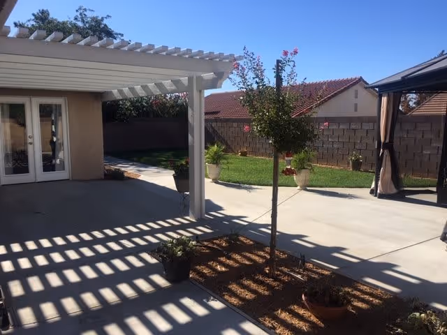 Backyard patio with a white pergola casting lattice shadows, a small planted tree, lawn, potted plants and a gazebo.