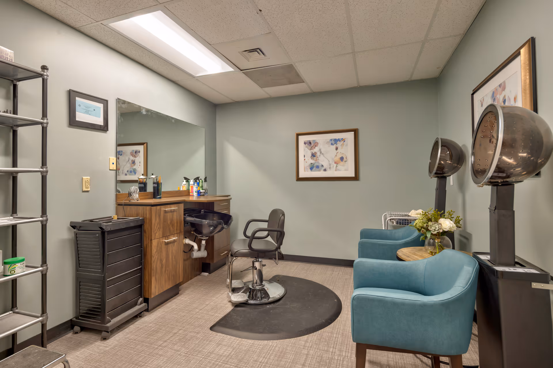 Interior view of a salon room with a styling chair in front of a large mirror, a black sink, and a wooden cabinet. Two blue armchairs are positioned near two vintage hair dryers. The room has light green walls, a framed abstract painting, and a metal shelving unit.