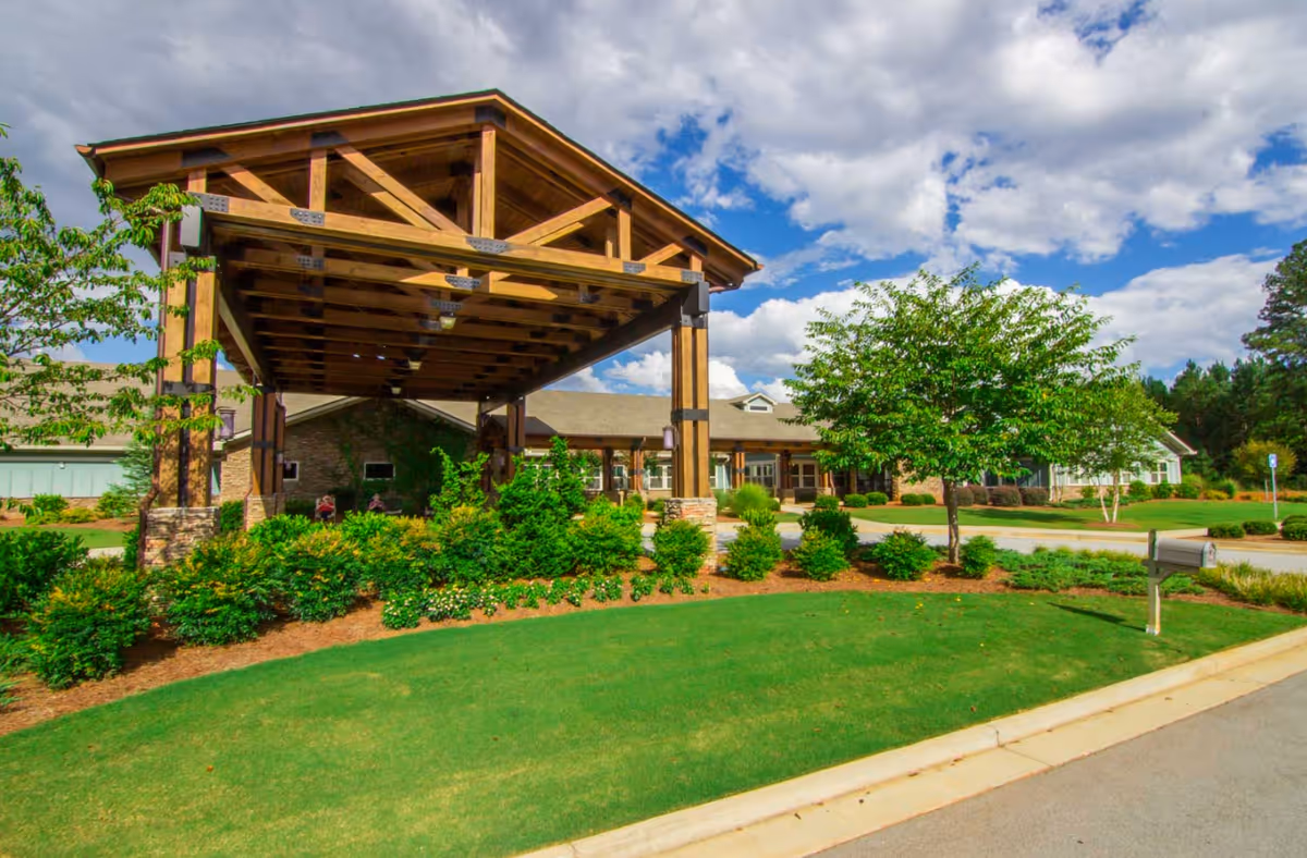 Exterior view of Oaks At Braselton facility showing a large wooden covered entrance with stone pillars, surrounded by green bushes, trees, and a well-maintained lawn under a partly cloudy blue sky.