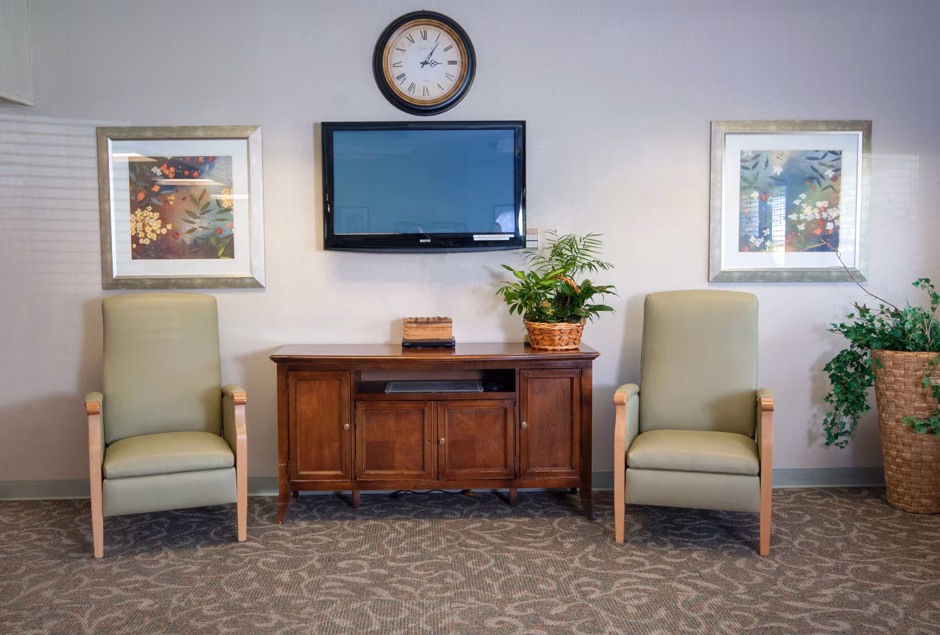 A sitting area with two light green armchairs on either side of a wooden cabinet. Above the cabinet is a wall-mounted flat-screen TV and a round clock. There are two framed floral paintings on the wall, one on each side of the TV. A potted plant sits on the cabinet, and another larger potted plant is on the floor to the right.