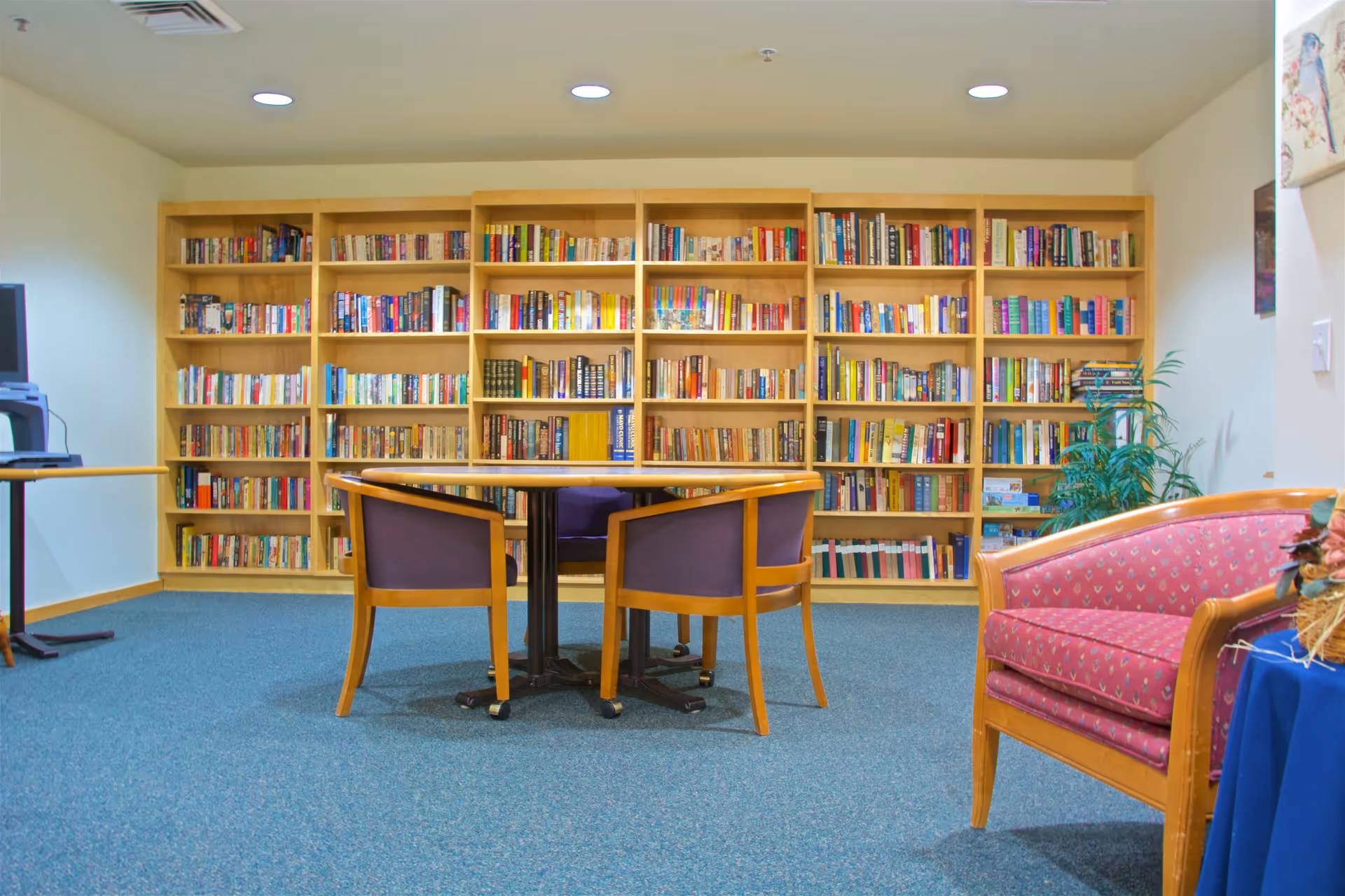A cozy reading room with a large wooden bookshelf filled with books along the back wall. In front of the bookshelf is a round table with three wooden chairs that have purple cushions. To the right, there is a wooden armchair with red patterned upholstery and a small table with a floral arrangement. The room has blue carpet and recessed ceiling lights.