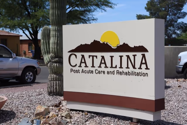 Outdoor sign for Catalina Post Acute Care and Rehabilitation with a mountain and sun logo, surrounded by desert landscaping including rocks and a large cactus, with parked vehicles and trees in the background.