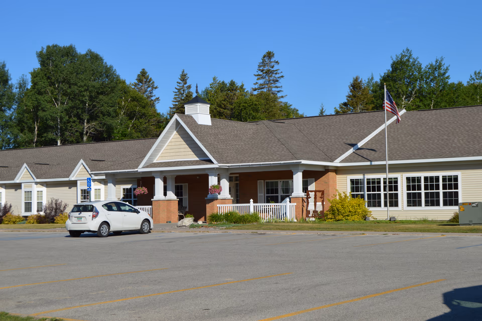 Exterior view of Cedar Cove Assisted Living facility showing a single-story building with beige siding, a brown shingled roof, white trim, and a covered entrance with columns. There is a white car parked in front near a handicap parking sign, an American flag on a flagpole, and trees in the background under a clear blue sky.