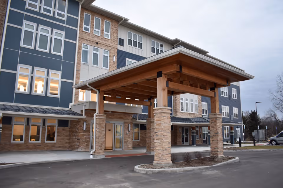 Exterior view of the entrance to Green Oaks of Goshen facility, featuring a covered drop-off area with wooden beams and stone pillars, a multi-story building with blue and beige siding, multiple windows, and a paved driveway.