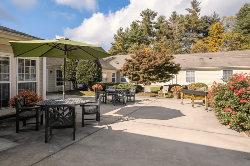 Outdoor patio area at Carolina Reserve of Hendersonville with several black tables and chairs, one table shaded by a large green umbrella. Surrounding the patio are bushes with red flowers, a small tree, and a beige building with multiple windows. The sky is partly cloudy with blue patches visible.