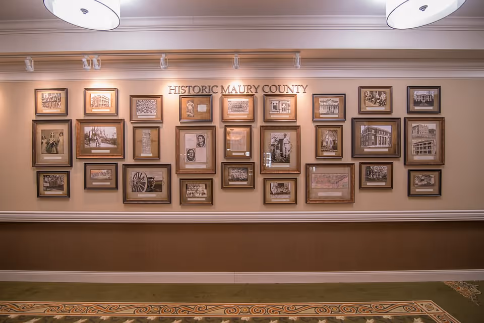 A wall display featuring multiple framed historical photographs and documents under the title 'HISTORIC MAURY COUNTY' in a well-lit interior space with beige walls and decorative carpet.
