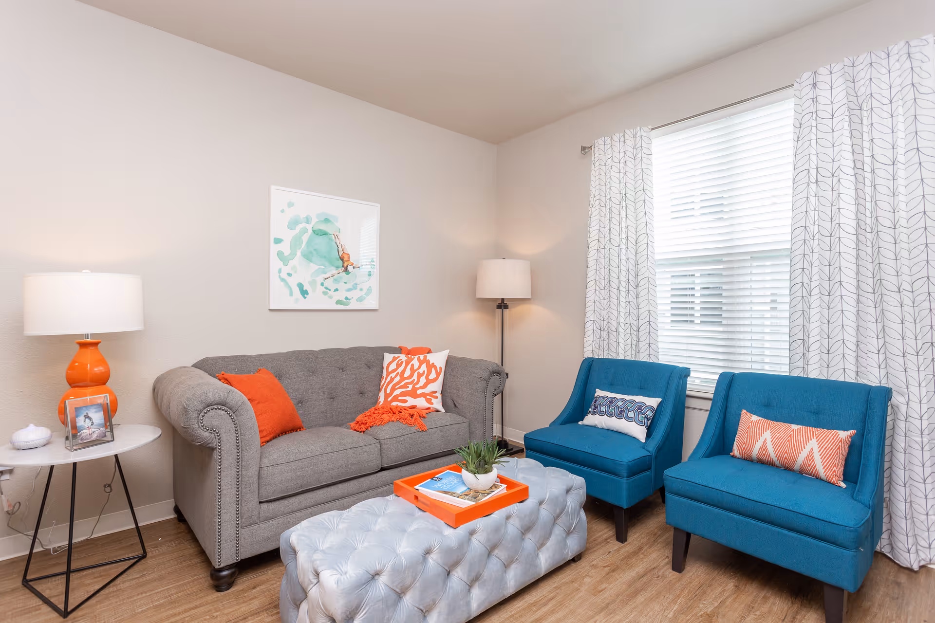A cozy living room with a gray tufted sofa adorned with orange and white pillows, two blue armchairs with decorative pillows, a large tufted ottoman serving as a coffee table with an orange tray and a small plant on it, a side table with an orange lamp and a framed photo, a floor lamp in the corner, and a window with white patterned curtains letting in natural light.