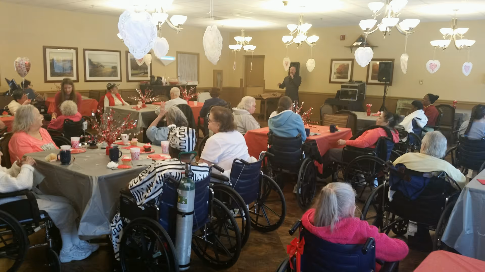 A group of elderly people, many in wheelchairs, seated around tables covered with red and gray tablecloths in a decorated dining room. The room has hanging heart-shaped decorations and chandeliers. A person is standing at the front of the room speaking or singing into a microphone.