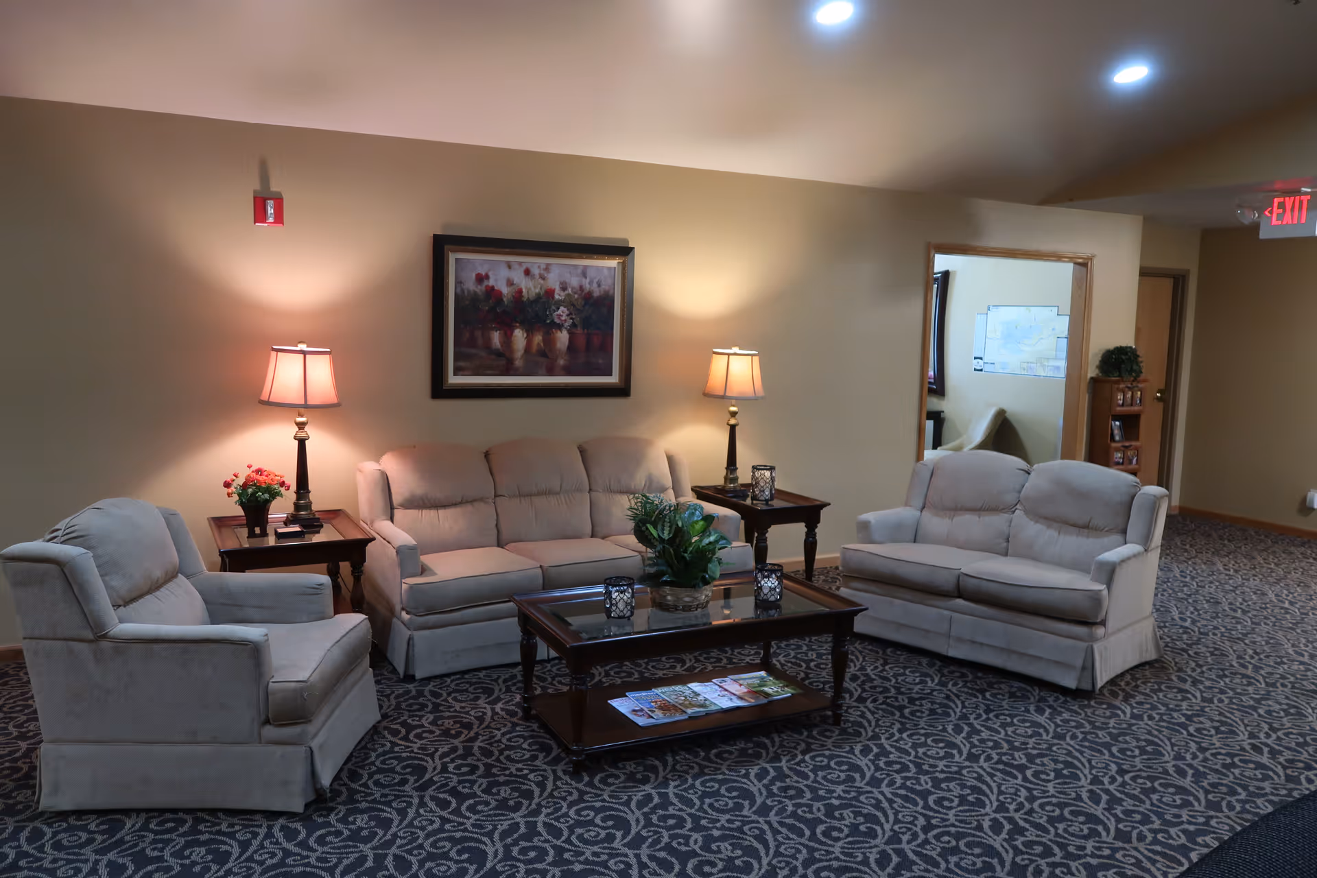 A cozy senior living facility common area with two beige sofas and one armchair arranged around a wooden coffee table with a glass top. The coffee table has a plant and some candles on it, and there are two side tables with lamps and a small flower pot. The walls are beige with a framed floral painting hanging above one sofa. The carpet has a dark pattern, and there is an open doorway leading to another room with a chair and a map on the wall.