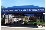 Entrance of Fairlane Senior Care & Rehab Center with a blue canopy displaying the facility's name. There are plants and shrubs along the walkway leading to the entrance.