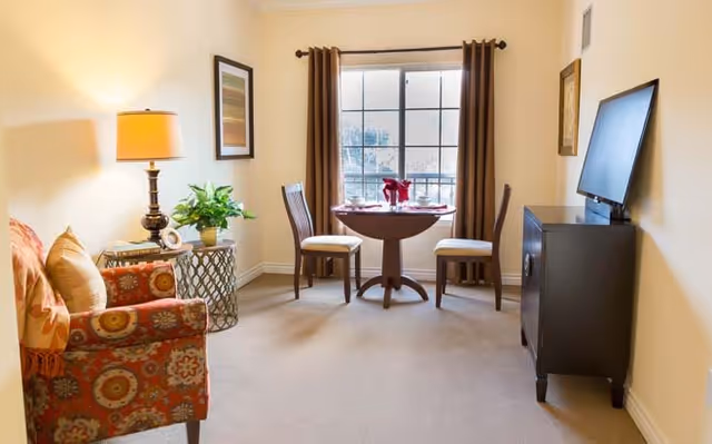 A cozy living room area with a patterned armchair, a side table with a lamp and a plant, a small round dining table set for two with red napkins, two chairs, a window with brown curtains, and a flat-screen TV on a dark wooden stand.