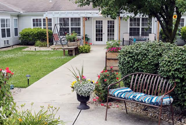 A landscaped courtyard with a paved walkway leading to a row of doors and windows, featuring benches, potted flowers, a swing, and shrubs.