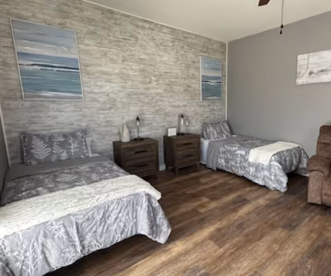 A senior living bedroom with two single beds, each with gray floral bedding and a white throw blanket. Between the beds are two dark wooden nightstands with lamps and decorative items. The walls are painted gray with one wall featuring a textured design and two framed ocean-themed paintings. The floor is wood, and there is a brown recliner chair partially visible on the right.